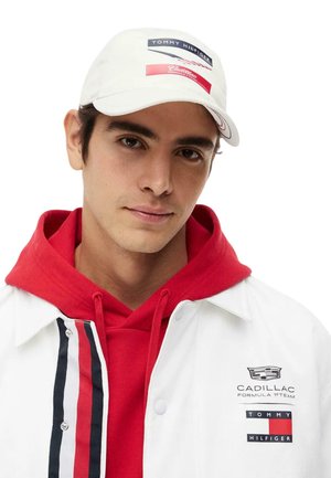 Giovane uomo che indossa un cappellino bianco Tommy Hilfiger e Cadillac, una felpa rossa con cappuccio e una giacca bianca con i loghi Cadillac e Tommy Hilfiger.