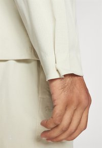 Light beige long-sleeve shirt with a textured pattern and buttoned cuff, paired with a matching cream-colored garment. Hand is visible.
