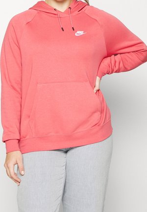 Personne portant un sweat à capuche Nike rose corail avec une poche avant et un pantalon gris clair, main sur la hanche, recadré sous le cou.