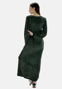 Długa, ciemnozielona, fakturowana sukienka z dzwonowymi rękawami i luźnym krojem, w połączeniu z białymi i czarnymi butami. Minimalistyczny design, bez wyraźnych wzorów.