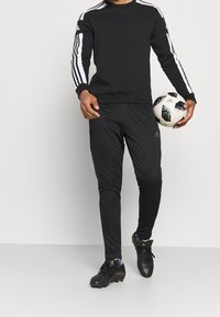 Svart idrottströja med vita ränder på ärmarna, ihopparad med svarta byxor och fotbollsskor, hållande en svart-vit fotboll.