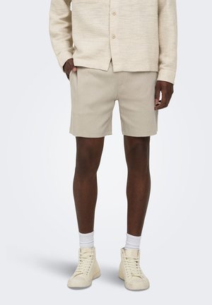 Homme portant un short beige texturé, une chemise boutonnée assortie, des chaussettes blanches et des baskets montantes crème, debout les mains dans les poches.