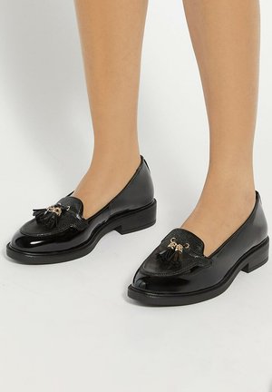 Black patent leather loafers with a textured panel and tassel detail, featuring a low block heel and rounded toe design.