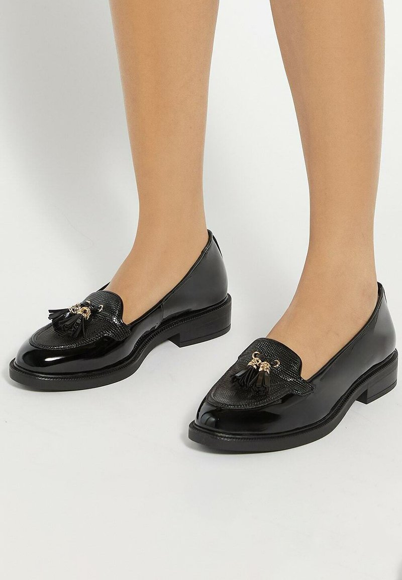 Black patent leather loafers with a textured panel and tassel detail, featuring a low block heel and rounded toe design.