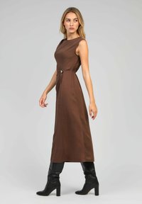 Robe longue marron avec un design sans manches, présentée avec un détail en torsion sur le côté et un accent doré rond à la taille. Portée avec des bottes noires jusqu'aux genoux.