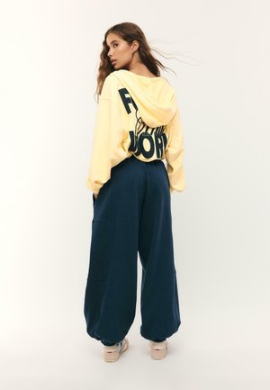 Femme aux cheveux longs portant un sweat à capuche jaune ample avec un texte en gras et un pantalon large bleu foncé, debout devant un fond clair uni.