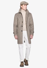 Brun houndstooth frakke med høj krave, fem knapper, parret med en beige turtleneck og hvide bukser. Brune sko fuldender outfittet.