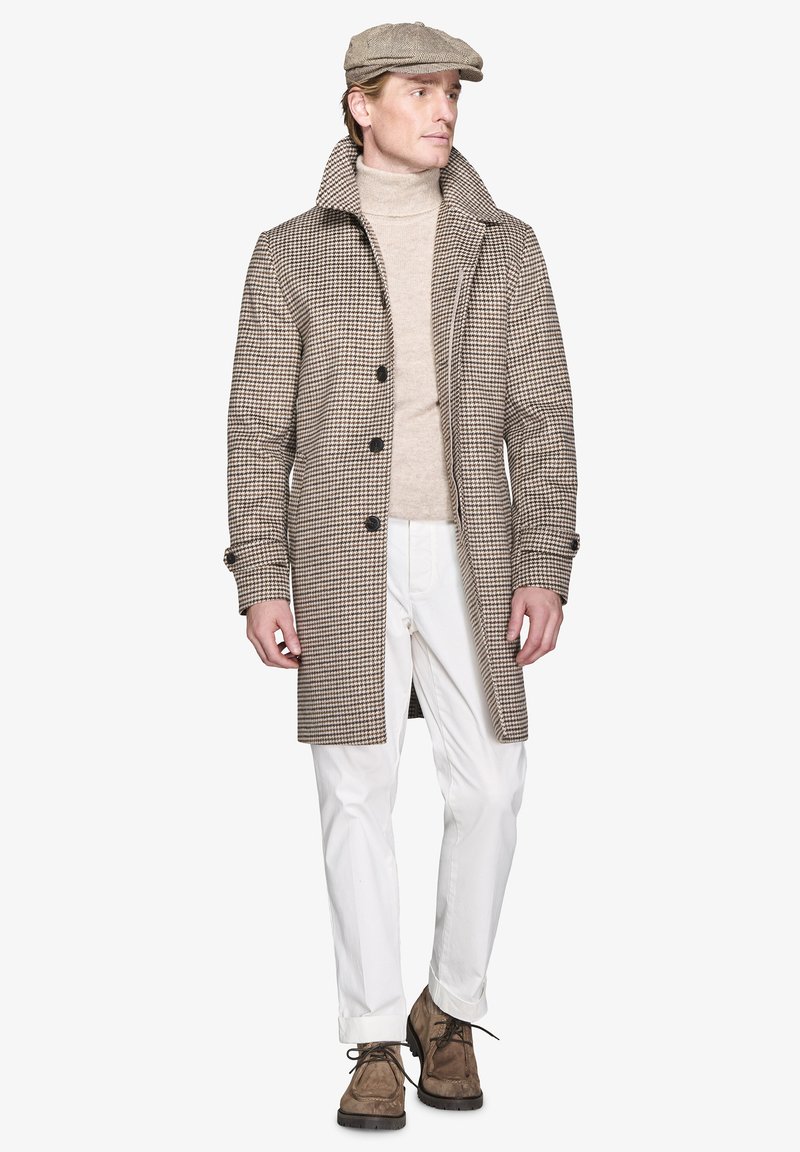 Brun houndstooth frakke med høj krave, fem knapper, parret med en beige turtleneck og hvide bukser. Brune sko fuldender outfittet.