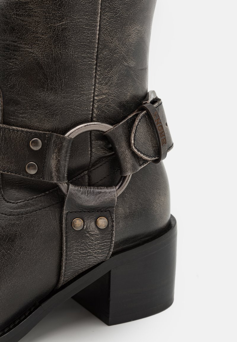 Stivale in pelle nera con tacco, dotato di una fascetta decorativa e dettagli in metallo a forma di anello, che evidenzia la texture e le cuciture su sfondo bianco.