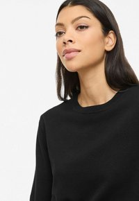 Mustast pehmest kootud materjalist kampsun, millel on ümar kaelus ja pikad varrukad, eksponeeritud neutraalse tausta ees.