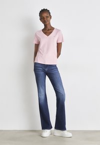 Camiseta de algodón rosa claro con cuello en V y mangas cortas, combinada con pantalones de mezclilla azul de corte acampanado y zapatillas blancas. Diseño sencillo, ajuste casual.