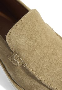 Suède instapper in lichtbeige met een ronde neus, verhoogde stiksels en een klein gespdetail aan de zijkant. Zacht textuur en casual ontwerp.