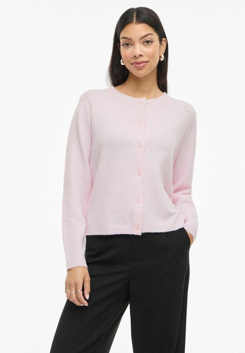 Rozā kardigans ar apaļu kakla izgriezumu, garām piedurknēm un priekšējiem pogu aizdares. Viegls adījuma materiāls un kraukšķīgs siluets, saskaņots ar melnām biksēm.