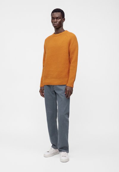 Homme debout portant un pull en tricot orange, un jean gris et des baskets blanches sur un fond clair uni.
