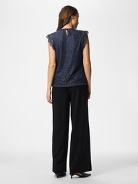 Nő sötétkék csipkés ujjatlan felsőben, hullámos szélű, fekete széles szárú nadrágban, sima háttér előtt áll, háttal a nézőnek.