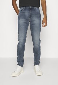 Jeans de mezclilla azul con corte slim, que cuentan con cinco bolsillos, pasadores para cinturón y un sutil patrón de desgaste; combinados con zapatillas blancas.