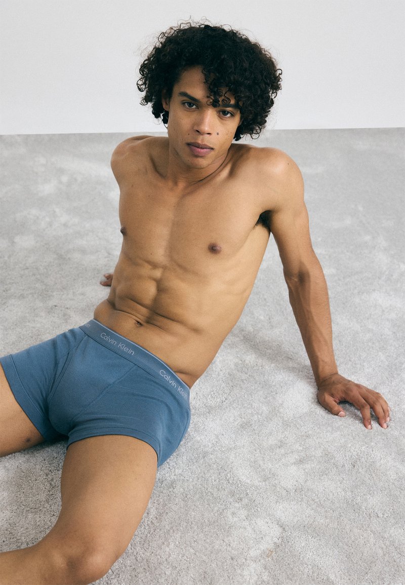 Boxerii scurți din bumbac albastru, cu o textură moale și o talie elastică pe care este înscris logo-ul 'Calvin Klein'. Modelul stă pe o suprafață gri deschis.