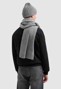 Grijze gebreide beanie en bijpassende sjaal, beide met ribbelstructuur. De outfit is gecombineerd met een zwarte trui en grijze jeans.