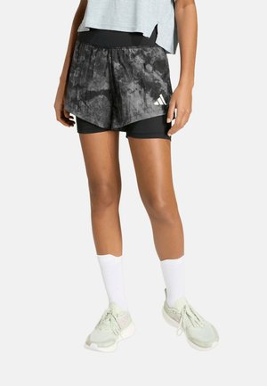 Persona que lleva puestos pantalones cortos de correr negros con estampado en capas, calcetines blancos hasta el tobillo y zapatillas de color claro, de pie frente a un fondo liso.