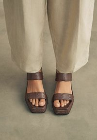 Brown, chunky sandals with wide straps, worn with light beige, loose-fitting trousers. The focus is on the feet against a muted background.