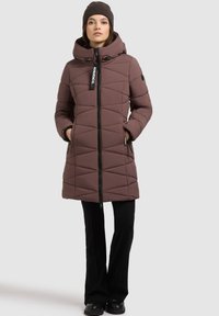 Bruine pufferjas met capuchon, voorzien van een ritssluiting aan de voorkant, een ruitpatroon en twee zijzakken, gecombineerd met zwarte broeken en een bijpassende beanie.