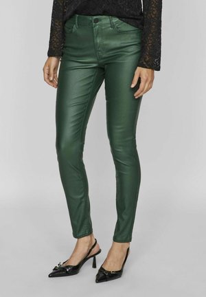 Femme portant un pantalon en cuir vert ajusté associé à un haut en dentelle noire à manches longues et des escarpins slingback noirs pointus avec un détail nœud.