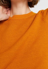 Orange stickad tröja med ett strukturerat mönster, med rund halsringning och något sänkta axlar. Närbild av tygdetaljer.