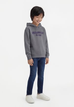 Ragazzo sorridente che indossa una felpa con cappuccio grigia DSQUARED2 Milano, jeans blu e sneakers bianche, in piedi contro uno sfondo bianco uniforme.