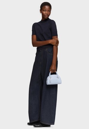 Mujer con una camiseta negra de manga corta y vaqueros de pierna ancha azul oscuro, sosteniendo un bolso pequeño de color azul claro sobre un fondo liso.