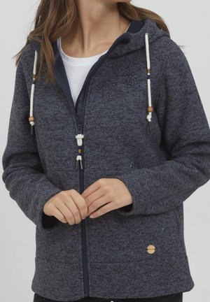 Kvinde iført en mørkeblå hættetrøje med lynlås og hvide snore med træperler over en hvid T-shirt, hænderne løst foldet ved taljen.