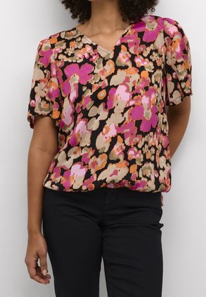 Femme portant un chemisier à manches courtes col en V avec un motif floral abstrait rose vif, orange, beige et noir, et un pantalon noir.