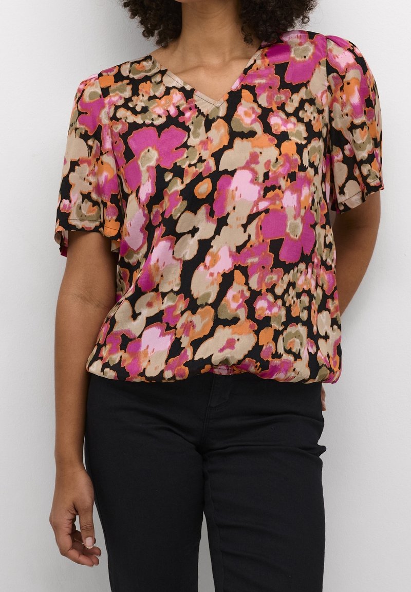 Femme portant un chemisier à manches courtes col en V avec un motif floral abstrait rose vif, orange, beige et noir, et un pantalon noir.