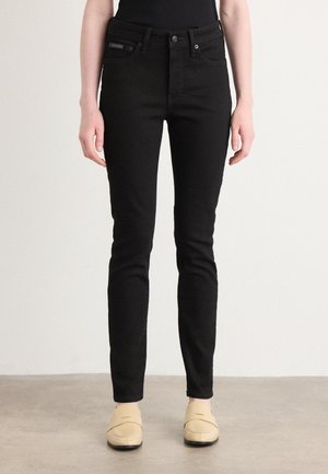 Kobieta w czarnych dopasowanych jeansach i beżowych wsuwanych butach, stojąca na jednolitym neutralnym tle.