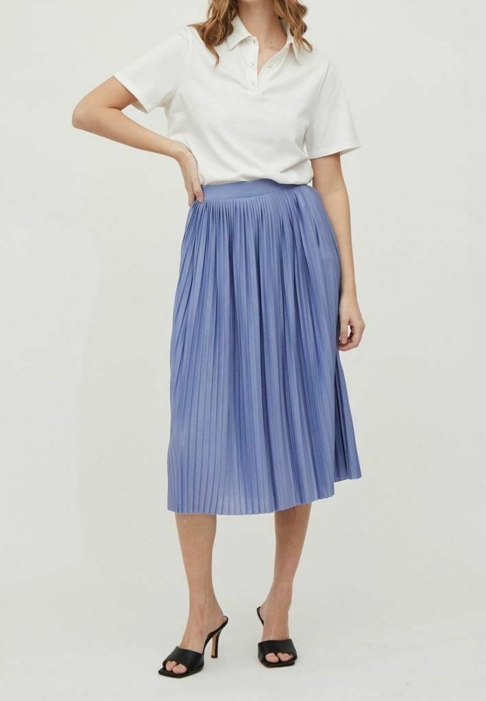 Femme portant un polo blanc à manches courtes, une jupe midi plissée bleue taille haute et des sandales à talons noires à bout ouvert.