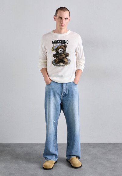 Vit tröja med björngrafik och "Moschino"-text, i kombination med ljusblå denimjeans och beige slip-on skor. Avslappnad passform, avslappnad stil.