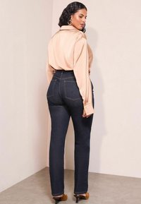 Blusa di chiffon pesca con maniche lunghe, abbinata a jeans a vita alta blu scuro e tacchi marroni, mostrata da dietro.