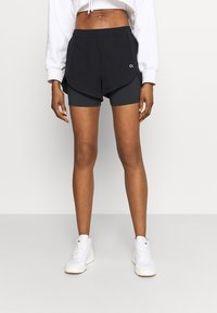 Pantalones cortos deportivos negros con capa interior, cintura elástica y diseño en capas. Combinados con zapatillas blancas sobre un fondo neutro.