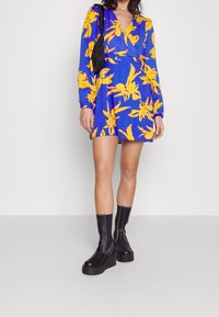 Robe bleue à motif floral jaune, manches longues et col en V. Portée avec des bottines à plateforme noires ornées de coutures blanches.