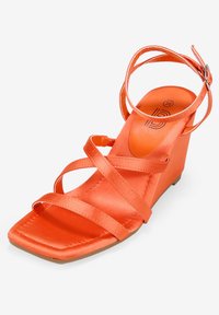 Sandalia naranja con tiras de satén, punta cuadrada y correa en el tobillo. Textura suave y diseño minimalista, con un tacón en cuña bajo.