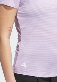 Fialová pruhovaná športová košeľa s krátkymi rukávmi a priliehavým strihom. Na spodnej ľavej strane sa nachádza biela logo. Koža je viditeľná s červeným tetovaním srdca.