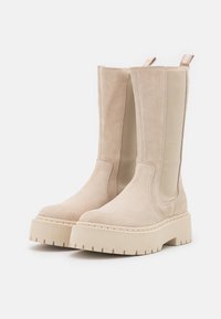 Bottes beiges en suede mi-mollet avec des panneaux élastiques sur les côtés, des semelles crantées épaisses et des tirettes à l'arrière sur un fond blanc.