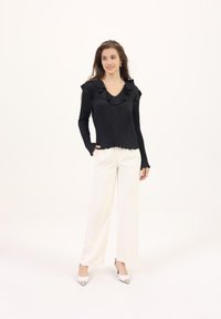 Black ribbed long-sleeve top with ruffled neckline, paired with white wide-leg trousers and white heels with silver accents, against a plain background.
