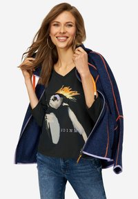 Giacca navy con cuciture arancioni indossata sopra una maglietta nera a scollo a V con una grafica di una scimmia. Il modello indossa jeans blu e grandi orecchini a cerchio.