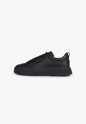 Zapatilla negra de perfil bajo con parte superior de cuero texturizado, suela gruesa acanalada, cordones delanteros y tirador en el lateral del talón sobre fondo blanco.