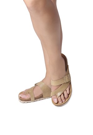 Beige sandals with cross straps, adjustable buckle, and textured footbed. Flat sole with slight heel and open-toe design.