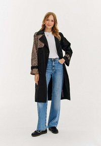 Čierny trenčkot s leopardími akcentmi na jednej strane, skombinovaný so svetlomodrými širokými nohavicami a čiernymi topánkami. Pod ním biele tričko.