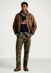 Brązowa skórzana kurtka na granatowym swetrze z wzorem amerykańskiej flagi, w połączeniu z kamuflażowymi spodniami cargo i brązowymi butami.