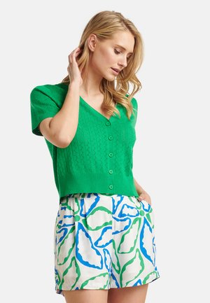 Green knitted cardigan with short sleeves, featuring a v-neck and button closure. Paired with white shorts, displaying blue and green abstract patterns.