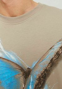 T-shirt in cotone beige con grafica di una farfalla con dettagli blu e marroni, scollatura rotonda e maniche corte. Texture morbida.