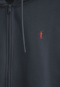 Granatowa bluza z zamkiem, wykonana z tkaniny o teksturowanym wykończeniu. Posiada czerwone wyszywane logo golfisty na lewej piersi.